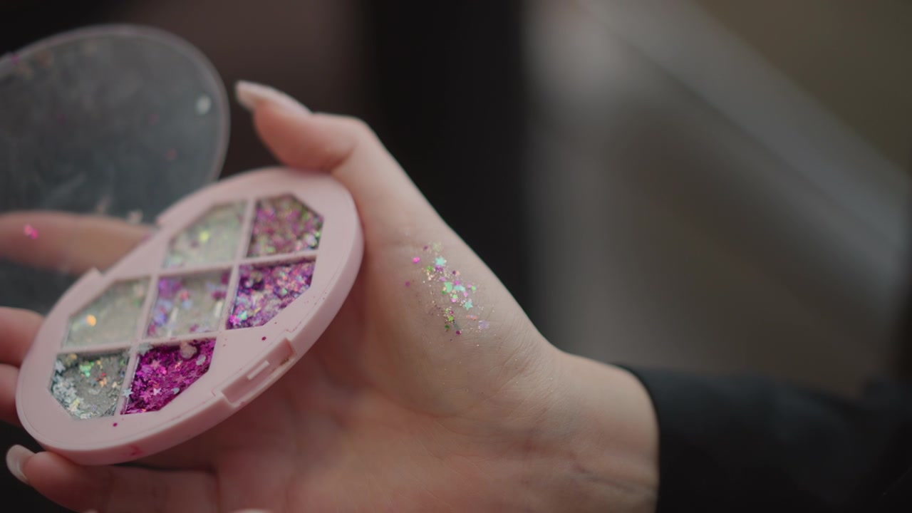 Close-up beauty scene showing hand holding round pink eyeshadow palette while loose glitter transfers onto skin and fingertips, with soft indoor light, pastel tones, reflective texture, and elegant