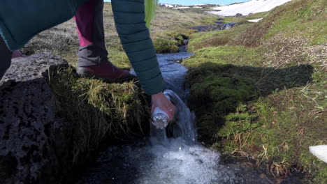 excursionista acercándose a un arroyo de montaña y llenando una botella de agua con agua dulce fría