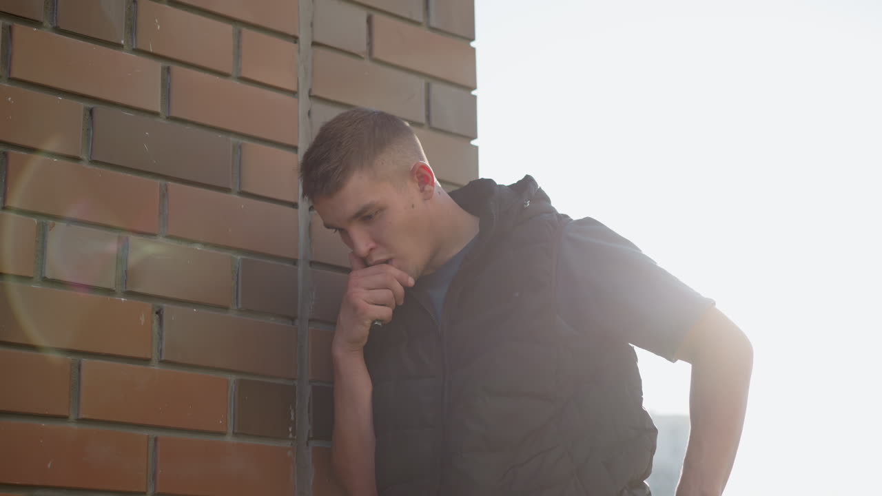 white man leaning on brick wall under sun flare coughing into hand after inhaling vape device emitting thick smoke wearing black puffer vest by urban apartment backdrop lens flare effect for moody