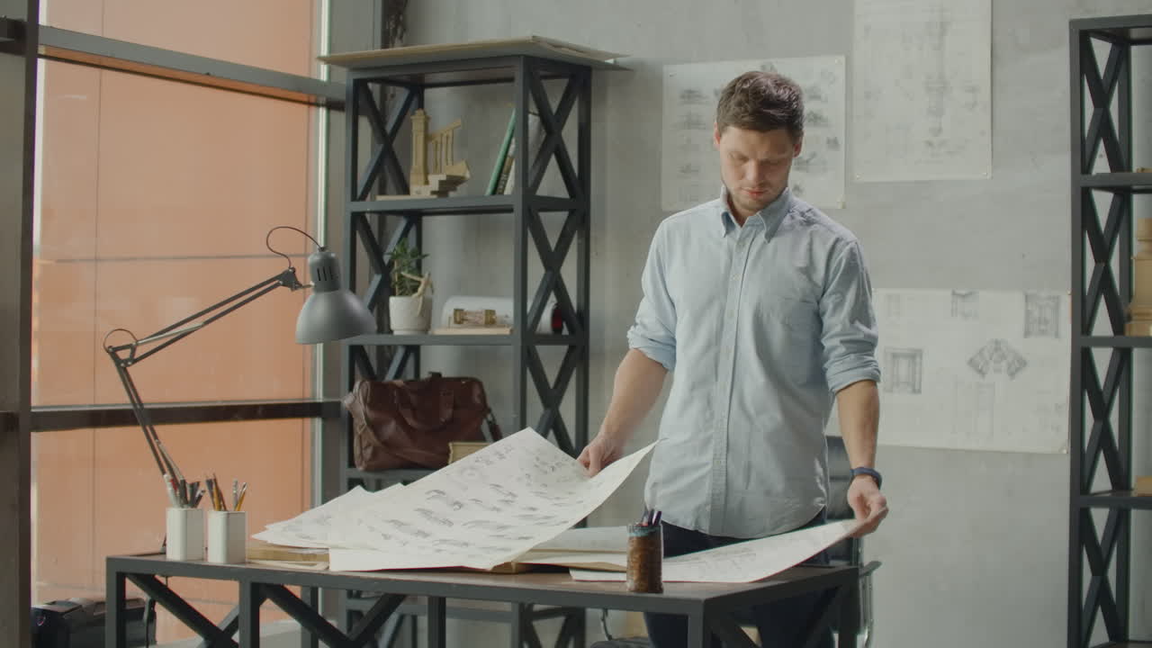 A male architect looks at drawings and plans of buildings in a loft office against a large window. The designer plans the facade of buildings. Creating furniture design