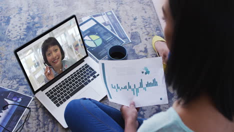african american woman holding a document having a video call with female colleague on laptop