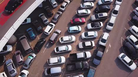 Vista-Aérea-Del-Vehículo-Que-Se-Mueve-Entre-Autos-Estacionados-En-Un-Estacionamiento-Abarrotado-Bajo-Una-Carretera-Elevada