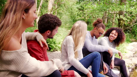 five young adult friends taking a break during a hike sitting on rocks by a stream in a forest, handheld