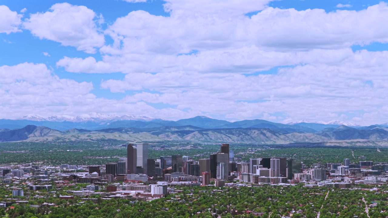 Et drone-luftfoto af Denvers bybillede, der viser skyskrabere og skyline mod de sneklædte Rocky Mountains i Front Range. Landskabet omkring downtown Denver, Colorado, med forårskvarterer under en blå morgenhimmel med skyer, panorering mod venstre.