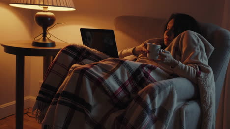 woman relaxing under a blanket with a laptop and a mug in a cozy, dimly lit room