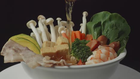 cerca de verter sopa en un cuenco de ingredientes de shabu girando alrededor de la mesa blanca en la cocina de fondo negro