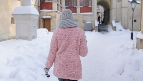 young woman in a pink coat and a hat drinking coffee on a snowy winter day in a city street