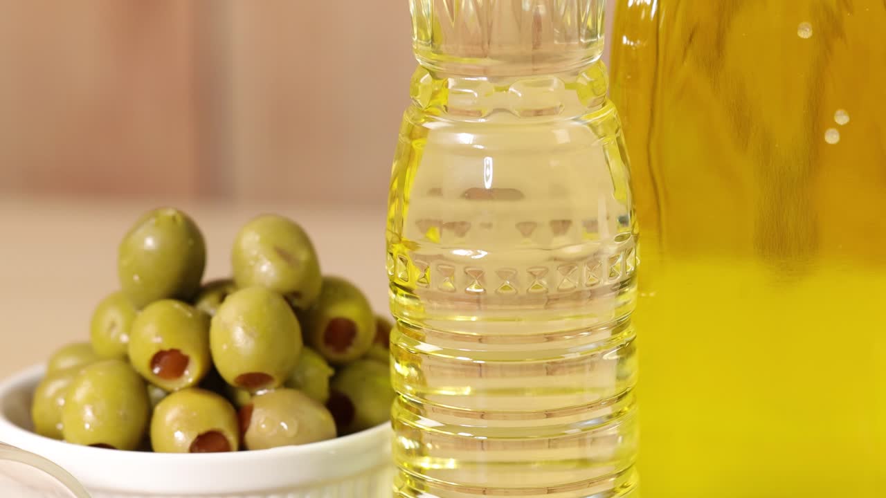Bottles of olive oil, a bowl of green olives, and a glass of almonds are arranged on a light wooden surface with soft, natural lighting and smooth camera movement