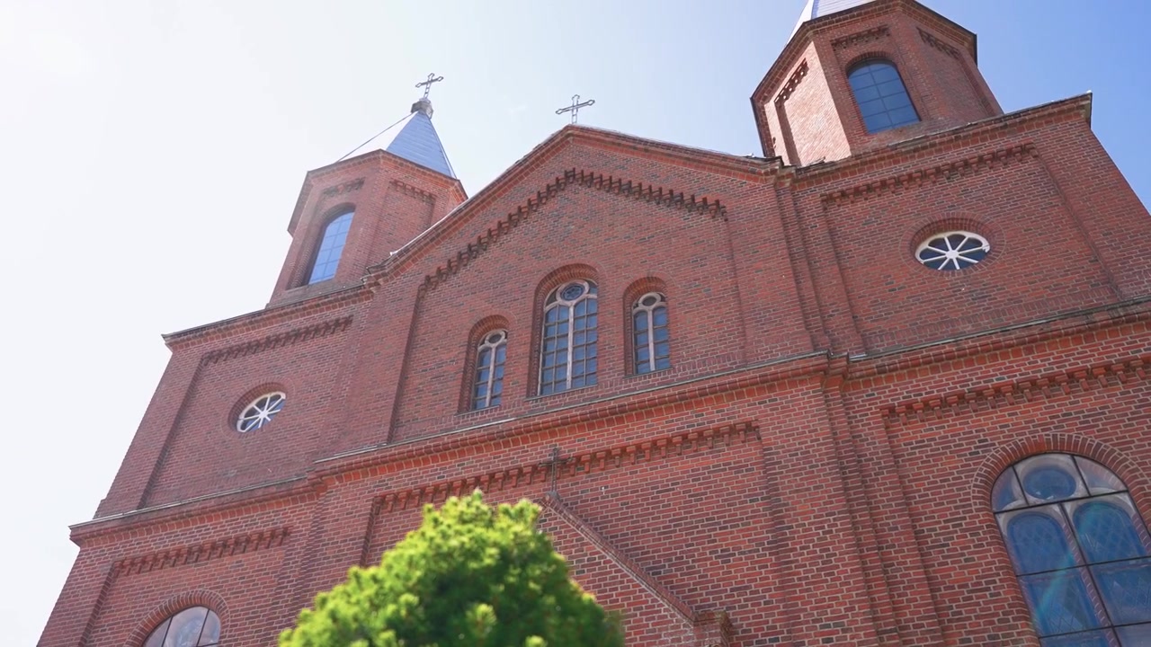 Tomada em ângulo baixo de uma impressionante fachada de igreja de tijolos vermelhos com torres gêmeas e janelas em arco, capturada sob luz do dia intensa. A luz do sol realça a arquitetura detalhada, a atmosfera vibrante e o céu azul claro.