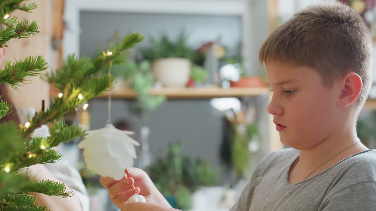 Close view of boy carefully fixing white flower ornament on christmas tree while mom assists in background, cozy indoor setting with festive decorations, preparing holiday atmosphere