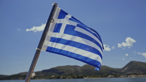 greek flag flying in the wind on boat with mediterranean island behind