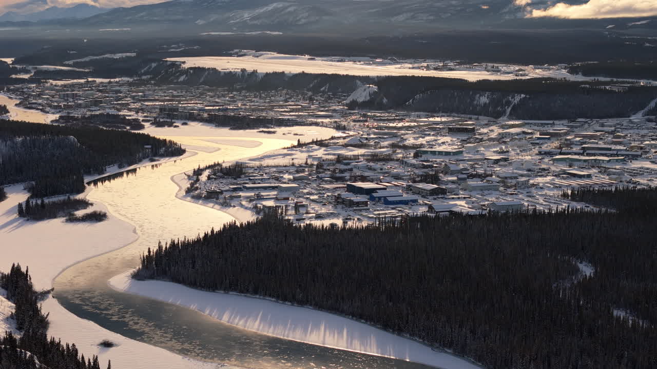 A cinematic aerial view of Whitehorse during golden hour. The snow-covered city and glistening Yukon River reflect the warm hues of sunset.