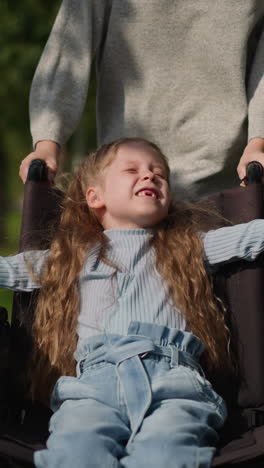 toothless little girl smiles in wheelchair spreading hands to sides. mother pushes medical vehicle with child running on road in spring park closeup