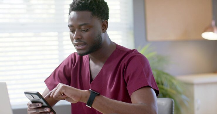 African American man tapping smartphone while speaking, glancing at laptop, reviewing notes at desk
