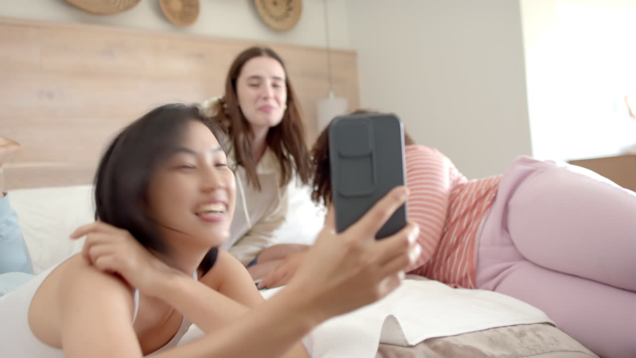 Taking selfie, three diverse women friends relaxing on bed and enjoying time together
