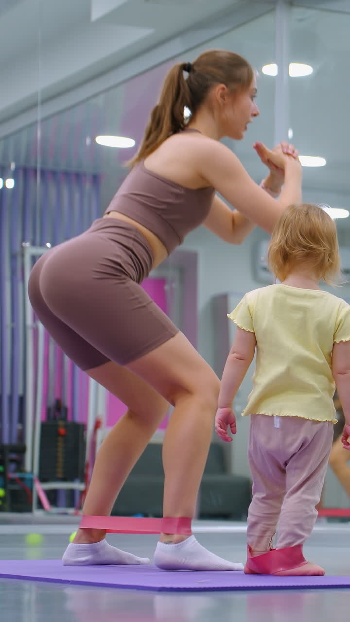 фитнес-сессия с участием женщины, сидящей на коленях с резистентной полосой, в то время как ребенок, также носящий резистентную полосу, наблюдает и имитирует ее движения, зеркала тренажерного зала отражают их действия