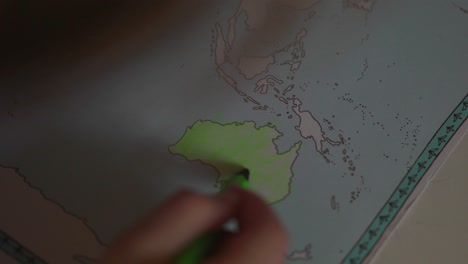 a toddler girl is coloring the continent of australia with a green pencil in a world atlas educational book