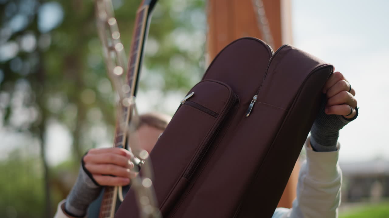 Giovane uomo bianco che apre una custodia per chitarra su un'altalena di legno in un parco illuminato dal sole, estraendo la custodia imbottita dalla catena, sollevando la chitarra acustica, regolando la tracolla, espressione concentrata, giacca di jeans, alberi sullo sfondo sfocato
