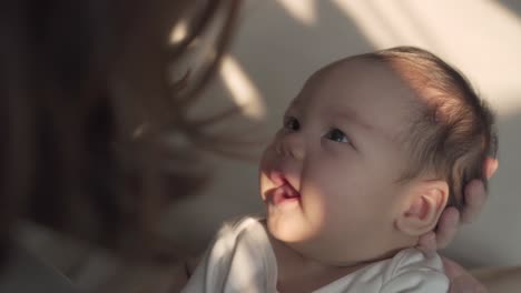 asian woman mother holding baby infant in arm playing and talking with kid on sofa couch in living room at home in evening. happy baby learning communication by smile, laugh, and stick out tongue.