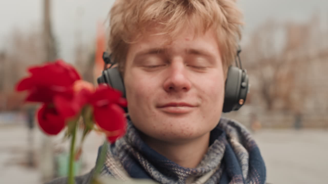 Young white creator on city sidewalk offering red rose, wearing headphones and scarf, autumn buses and muted buildings in background, hopeful expression and gentle smile, cinematic city portrait