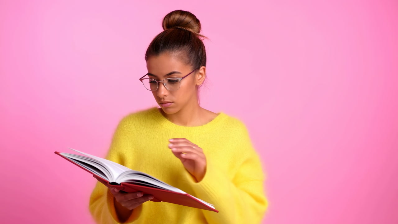 Young Woman Reading Book Against Pink Background, Yellow Sweater And Glasses, Hair In Bun, Turning Pages
