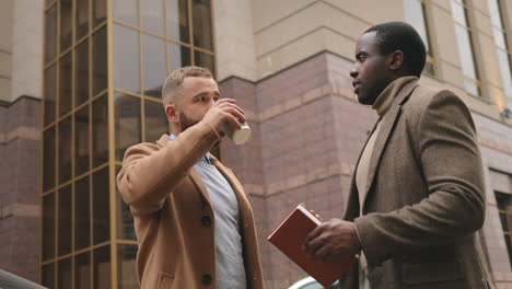 vista inferior de un hombre de negocios caucásico y afroamericano vestido con ropa elegante hablando en la calle en otoño mientras uno de ellos bebe café y el otro sostiene un smartphone