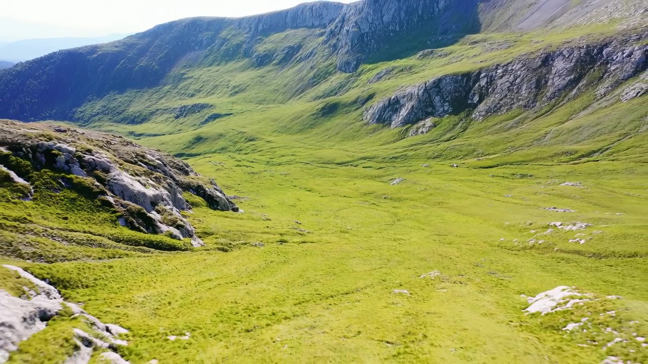 Ampia valle alpina mostra prati color smeraldo, rocce aspre, creste distanti, pendii ondulati, cielo azzurro limpido e luce diurna brillante attraverso un paesaggio naturale pacifico con orizzonte aperto, con elementi alpini e vallivi