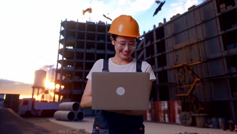 trabajadora asiática con gafas de protección y casco de seguridad usando una computadora portátil mientras está de pie en el sitio de construcción