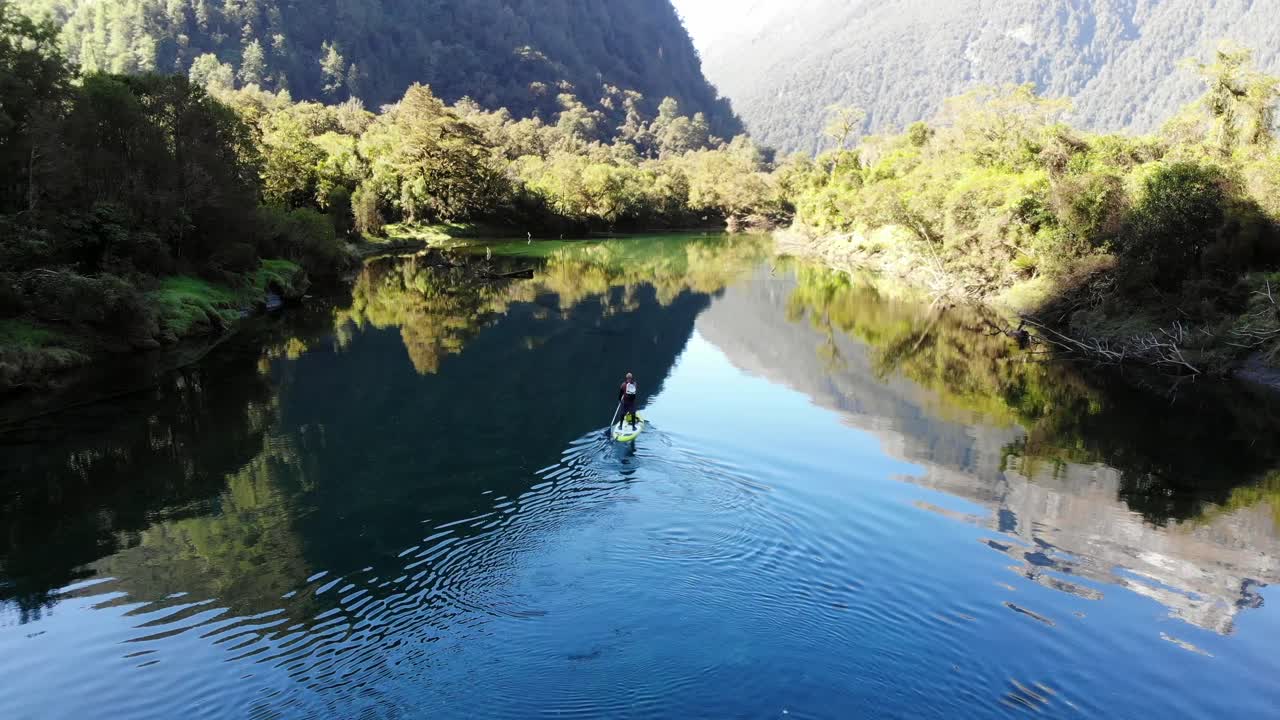 Man paddle boarding down stunning mountain river with crystal clear water surrounded by rainforest and steep walled mountains