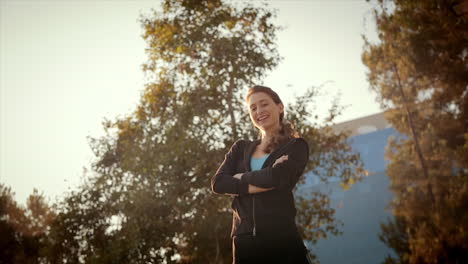 Low-angle-medium-dolly-shot-of-young-beautiful-slim-caucasian-woman-in-her-jogging-outfit,-crossing-her-arms-with-confidence-and-smiling,-laughing-at-the-camera