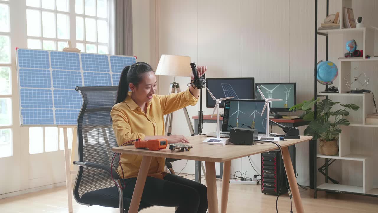 mujer asiática probando la turbina eólica mientras trabaja con una computadora portátil junto a la célula solar en la oficina