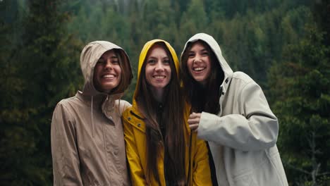 three girls in light jackets in hoods rejoice in the coming rain and look at the camera and hug in a mountain forest