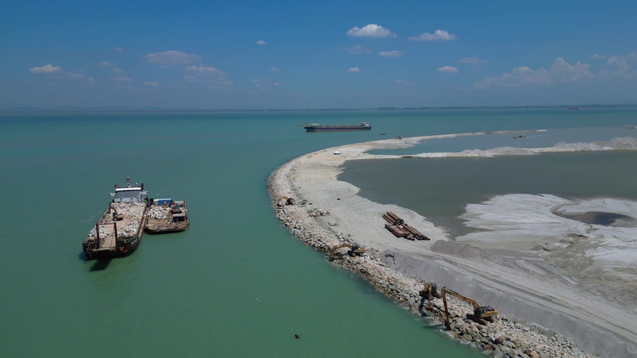 Cargo ships moving construction materials along a coastline with land reclamation work under a clear blue sky in Penang, Malaysia. Dramatic aerial view static panorama hovering drone