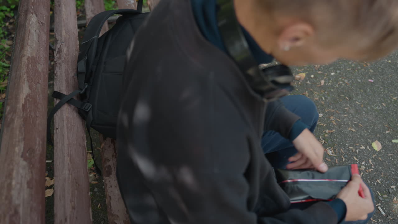 Junger Mann auf einer Parkbank, der eine tragbare Konsole spielt, Kopfhörer um den Hals, Rucksack an verwitterten Holzlatten lehnend, Laub und Kies unter den Füßen, lässige Jacke und Turnschuhe, konzentrierte Daumen