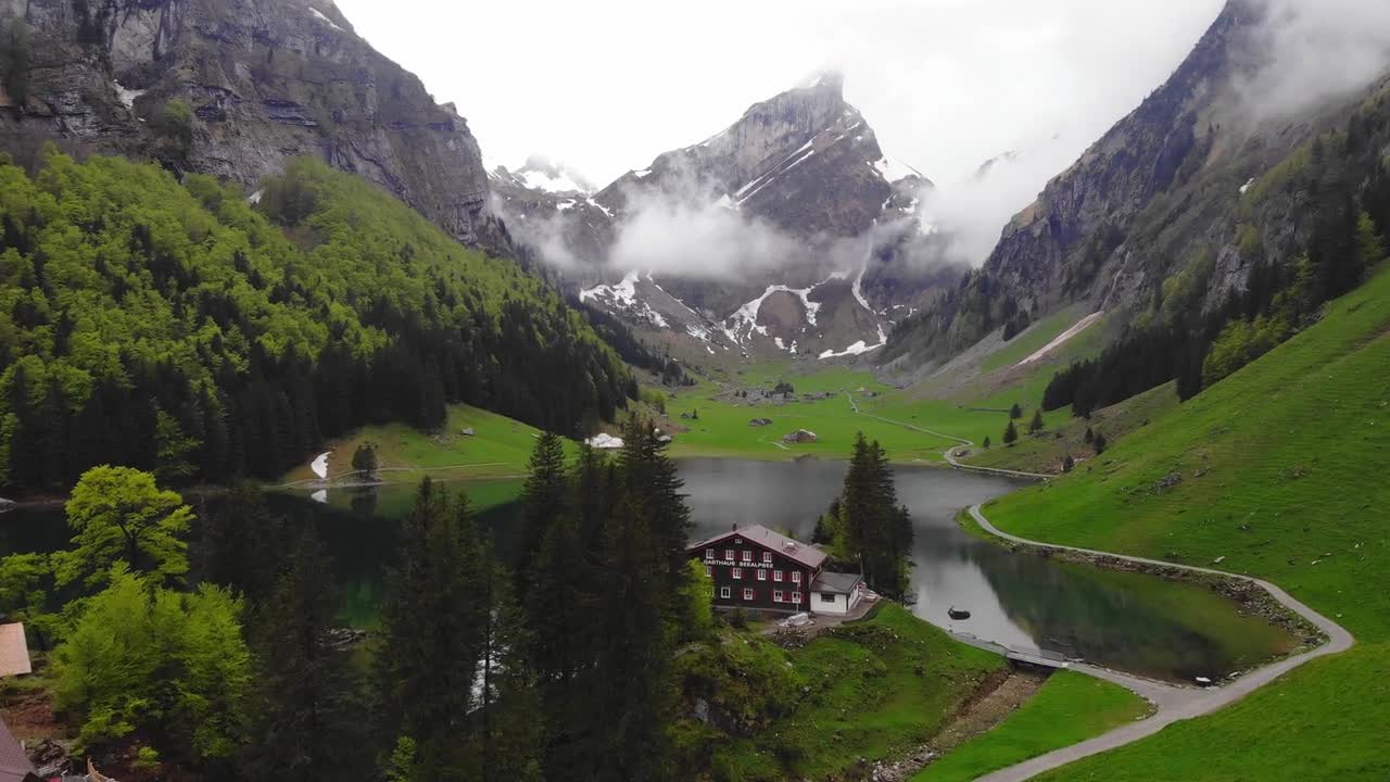 Drone captures picturesque lake and houses surrounded by towering mountain ranges in Iceland. The crystal-clear lake reflects the majestic peaks with lush greenery