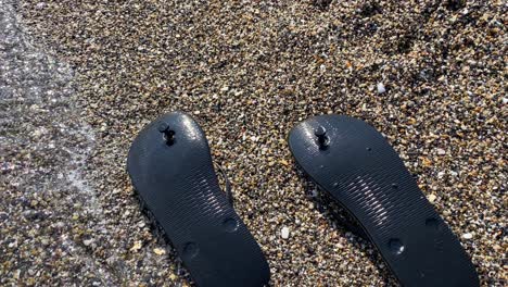 Wellen-Treffen-Auf-Angeschwemmte-Schwarze,-Umgedrehte-Flip-Flops-An-Einem-Strand-Mit-Winzigen-Kleinen-Felsen-In-Spanien,-Sonnige-Sommerferienschuhe,-4K-Aufnahme