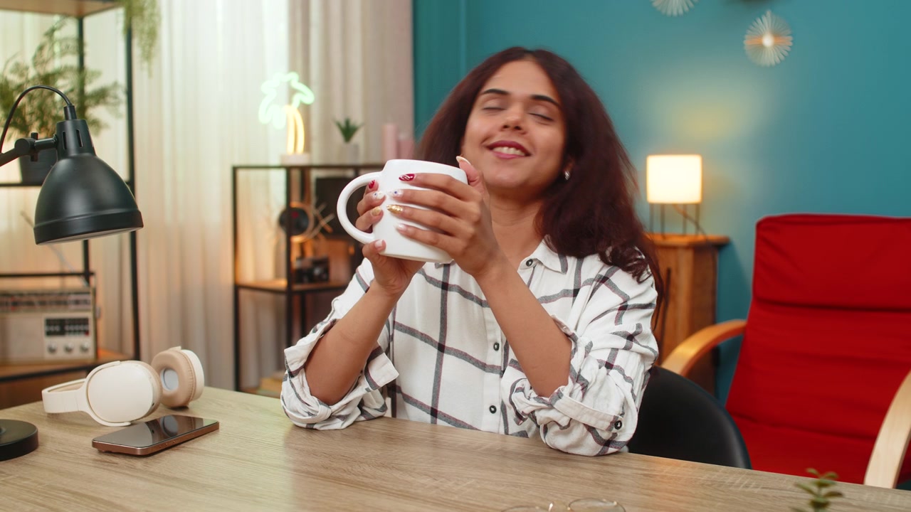 Mujer india disfrutando de un café caliente con los ojos cerrados, sosteniendo la taza, saboreando el aroma y relajándose tranquilamente en el interior