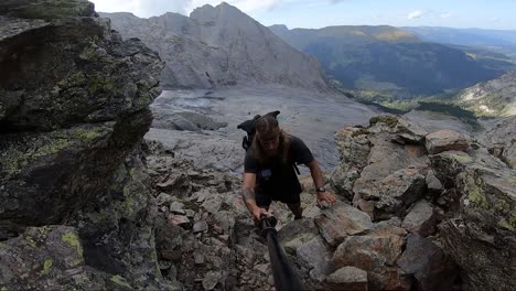 a young, strong and fit man with long hair and tattoos is hiking with his backpack up between rocks and cliffs on his way up to the peak of the mountain