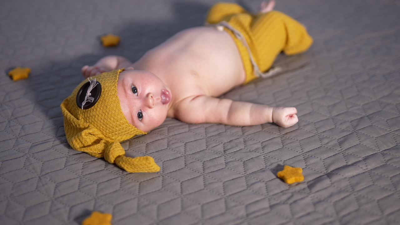 Cute baby boy in a yellow costume of a gnome lies on the bed. Decorative stars around the baby on the bed. Baby sticks out tongue.