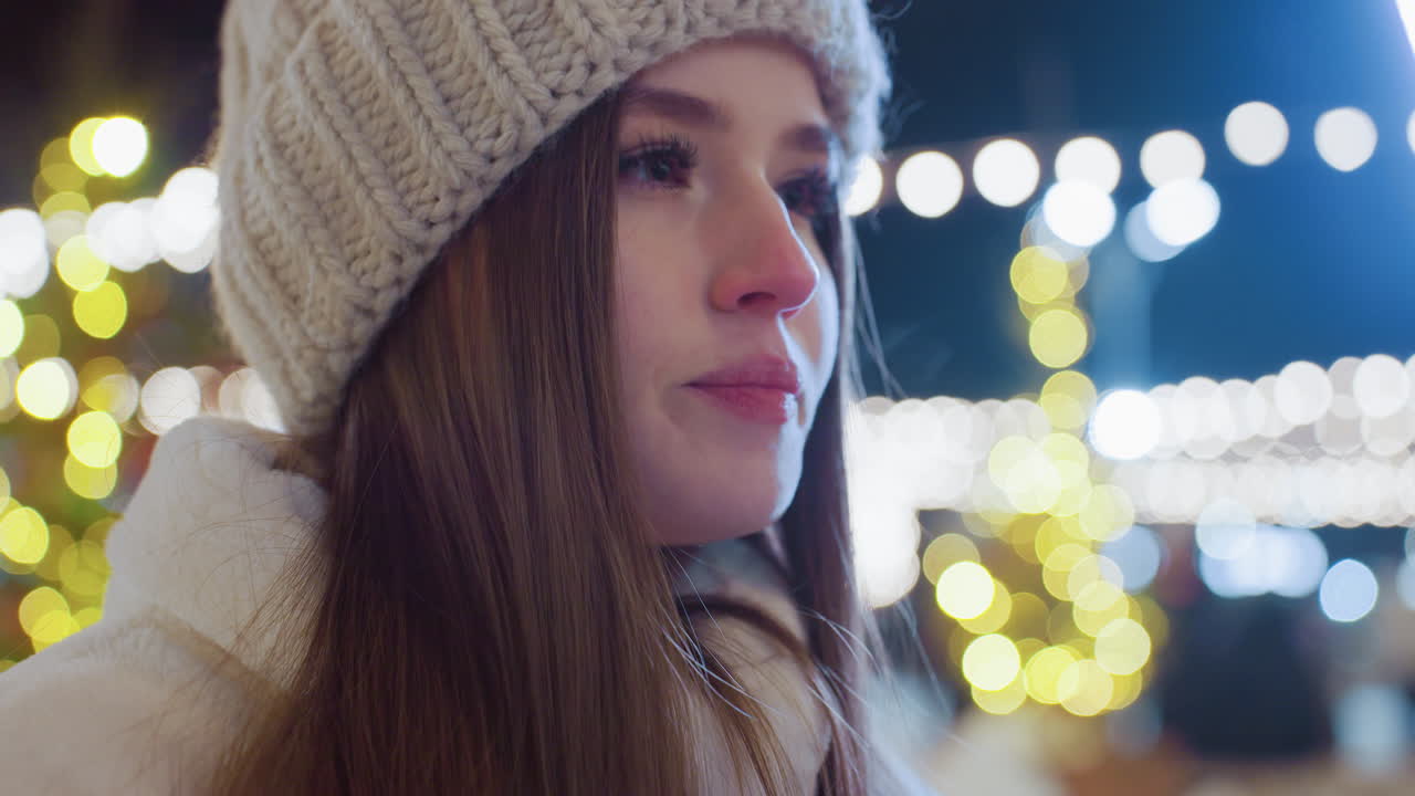 Close-up of young lady in warm winter outfit covering her mouth with gloved hands as visible cold breath escapes, festive city lights glow softly in the blurred background
