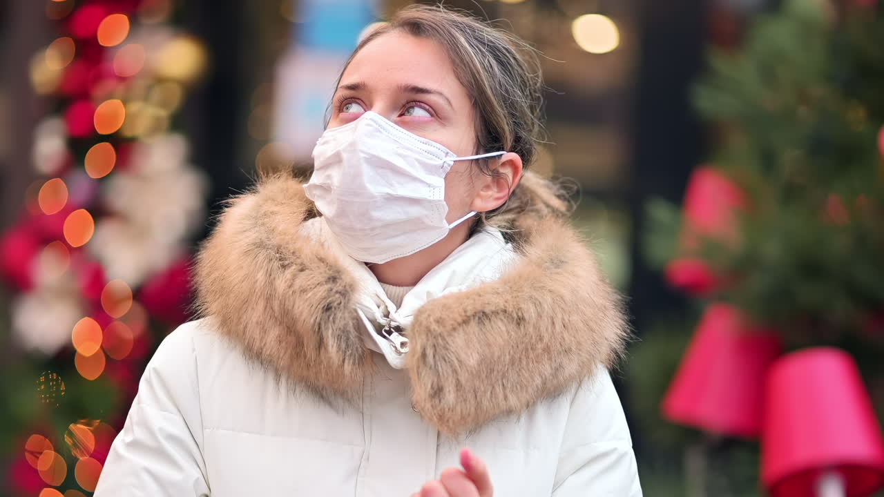 A young woman in a stylish white coat and face mask gazes at vibrant holiday decorations and lights in a bustling city street, capturing the joy of the festive season