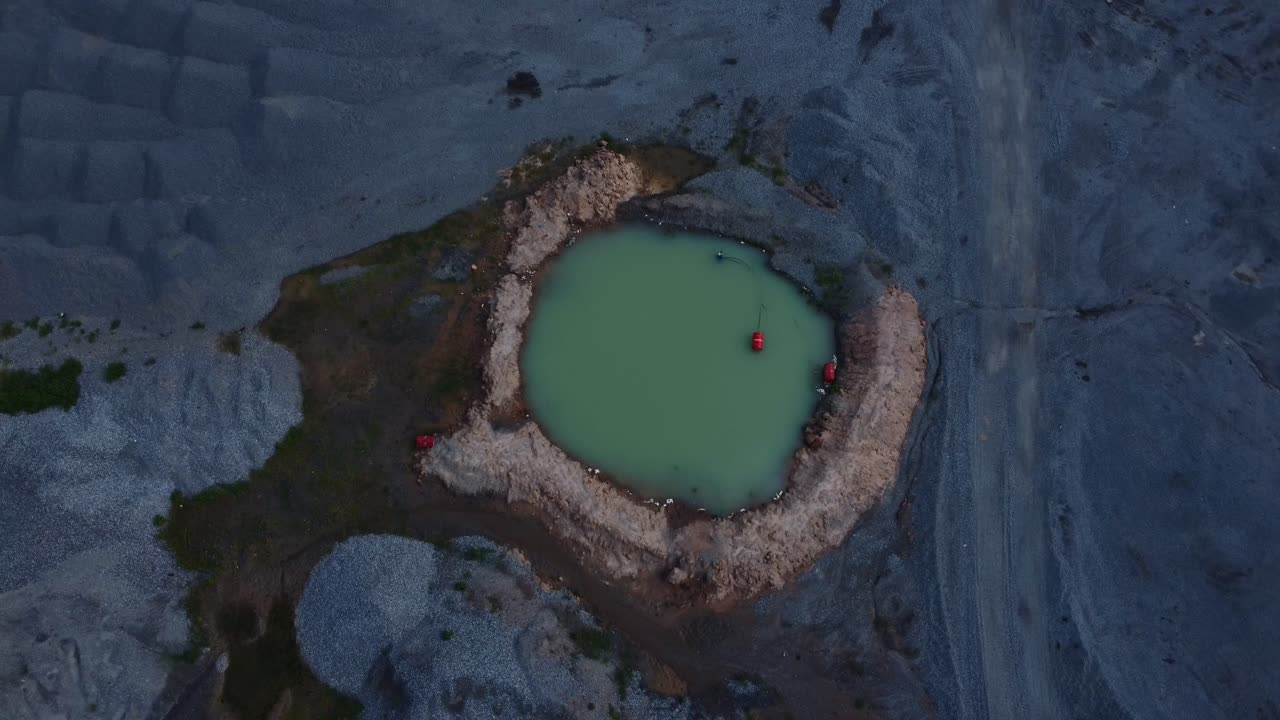 Aerial view of a solitary water pit in a rugged quarry setting at twilight. The scene captures the stark contrast of rocky terrain against the calm, green water, creating a tranquil industrial mood