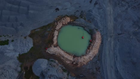 aerial view of a solitary water pit in a rugged quarry setting at twilight. the scene captures the stark contrast of rocky terrain against the calm, green water, creating a tranquil industrial mood