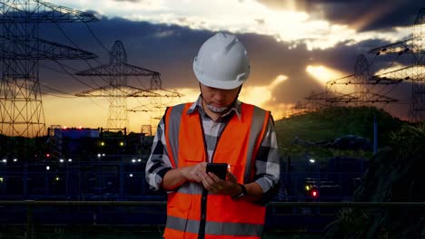 asian male engineer with safety helmet using smartphone while standing near high voltage tower