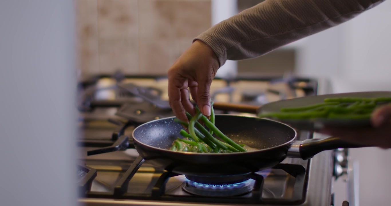 Hands are placing plate-held green beans flat into sizzling pan over blue gas flame to cook