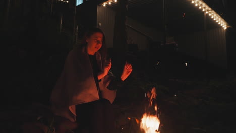 lady enjoys campfire warmth sitting nearby. young married woman with shoulders covered with blanket extends her hands over bonfire embracing evening glow