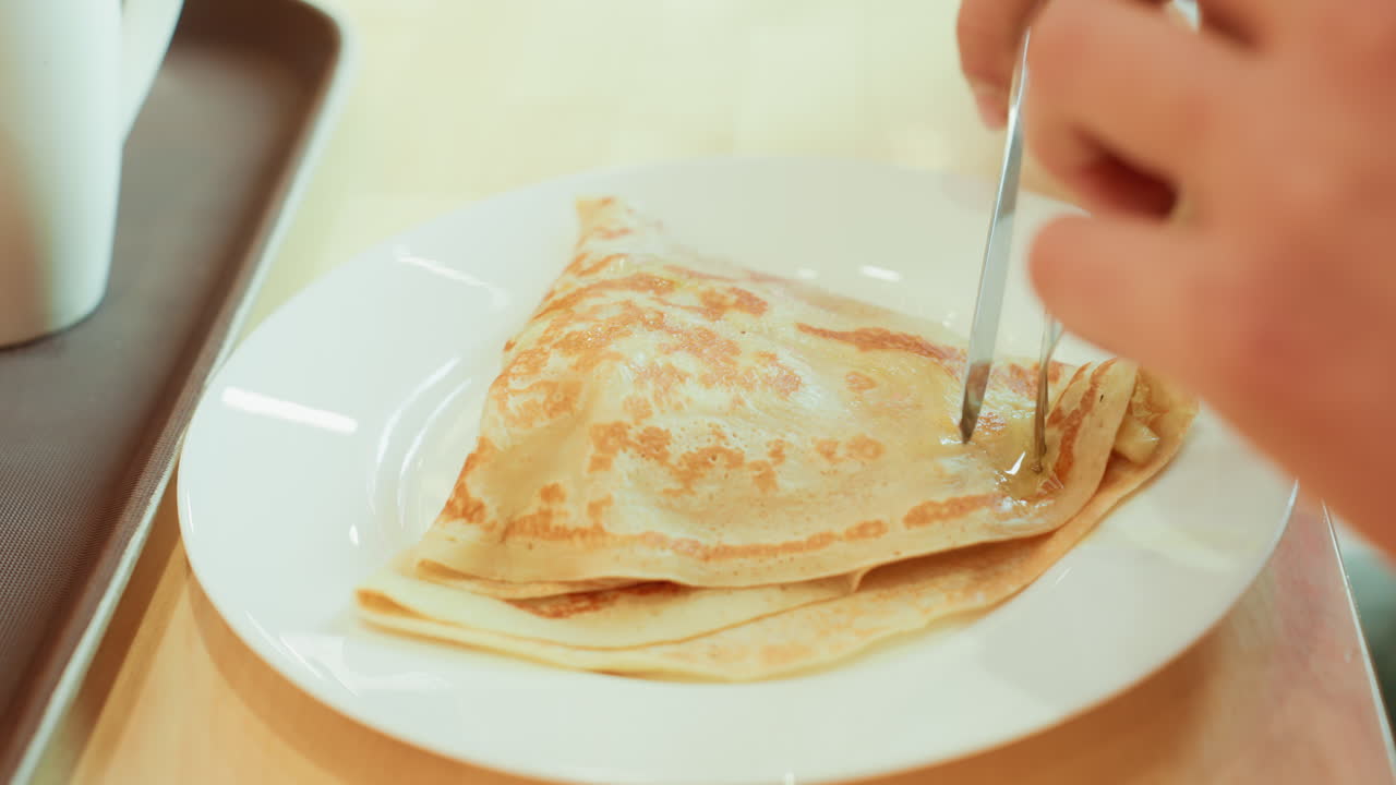 Mano che taglia una crepe a colazione. Crepe calda tagliata sul tavolo. Crepe fatta in casa servita con una delicata luce mattutina. Momento accogliente della colazione con mani che tagliano una crepe calda e zuccherata su una superficie di legno