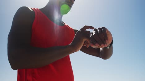 African-american-man-checking-his-smartwatch,-taking-break-in-exercise-outdoors-by-the-sea