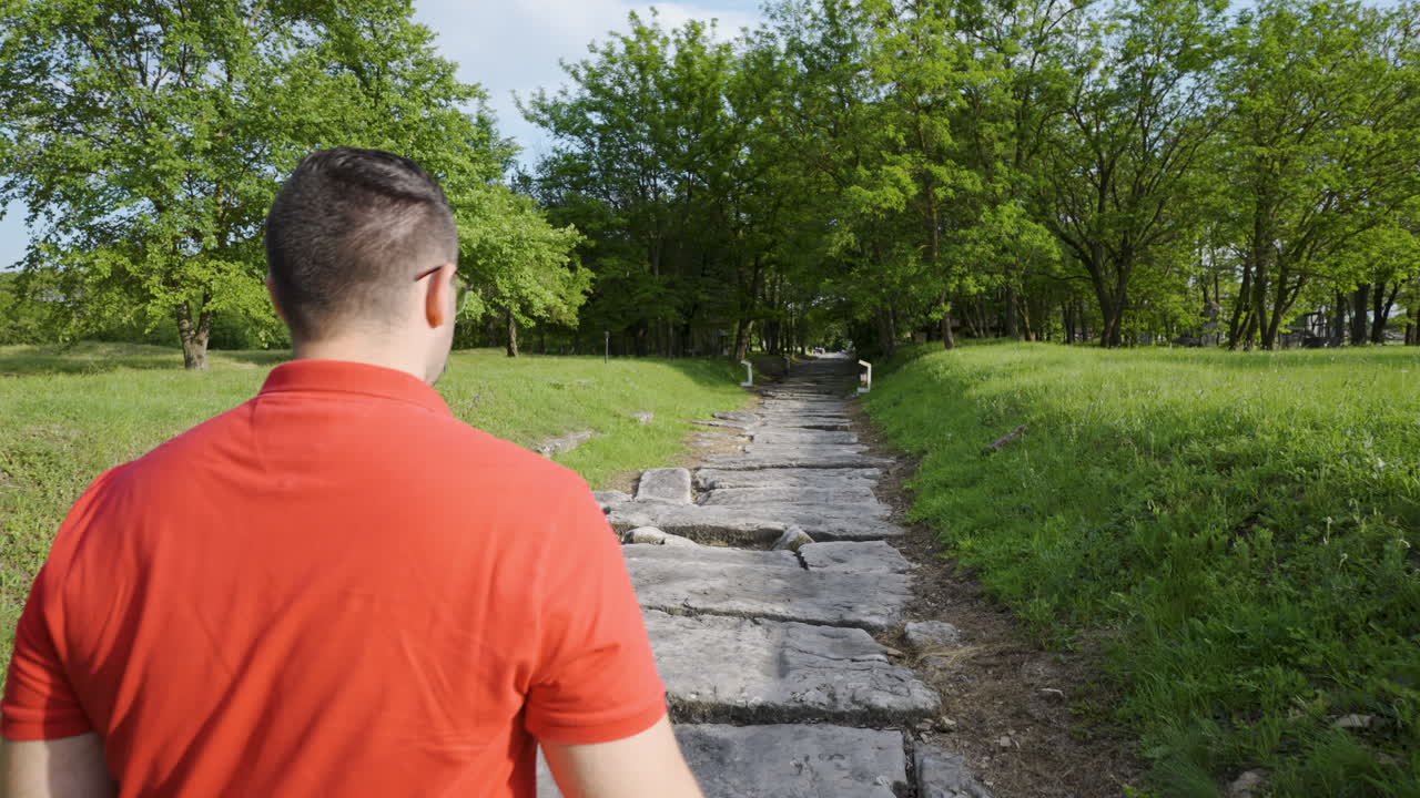 Follow shot of a man in a red shirt walking along a stone path surrounded by lush greenery during golden hour