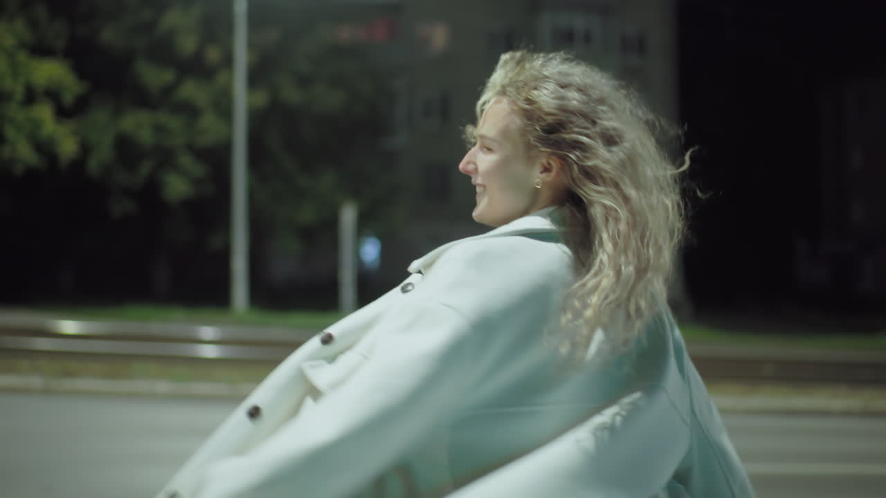 White girl exuding pure happiness as bright street light illuminates her smiling face while blurred residential building across road creates vibrant urban nighttime backdrop